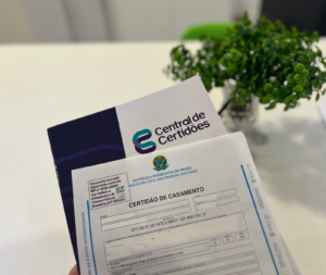 Imagem de uma certidão de casamento sendo segurada junto a um envelope da Central de Certidões, com uma planta ao fundo.