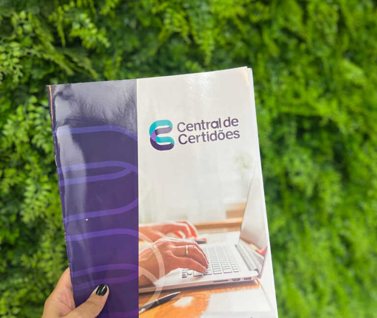 Folder da Central de Certidões segurado por uma mão, tendo como fundo uma parede verde de plantas.